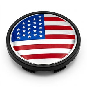 May include: A circular wheel center cap featuring a glossy American flag design with a blue field of white stars and alternating red and white stripes. The emblem is encased in a black plastic housing with a textured outer rim and mounting clips.