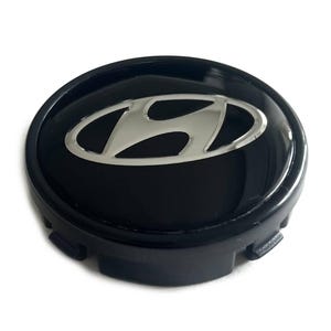 May include: A circular black wheel center cap featuring a silver Hyundai logo in the center. The cap has a glossy finish on the top surface and includes integrated plastic clips along the outer edge for secure attachment to a vehicle wheel.