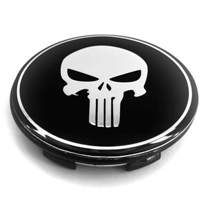 Könnte beinhalten: Eine kreisförmige Radnabenabdeckung mit glänzend schwarzer Oberfläche und einem markanten, silberfarbenen Totenkopf-Emblem in der Mitte. Das Emblem ist von einem dünnen silbernen Kreisrand umgeben, und die Unterseite weist schwarze Kunststoffclips auf.