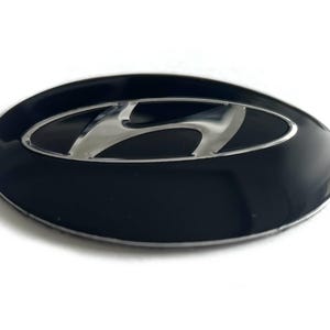 May include: An oval-shaped Hyundai vehicle emblem featuring a glossy black background with a raised, polished silver-tone metallic 'H' logo centered within the frame. The emblem has a smooth, reflective surface and a curved, three-dimensional profile.