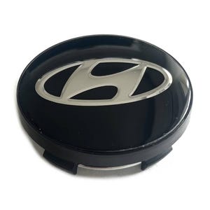 May include: A circular black wheel center cap featuring a silver-toned Hyundai logo emblem centered on the glossy surface. The cap has a matte black outer edge with integrated mounting clips visible along the bottom perimeter of the component.