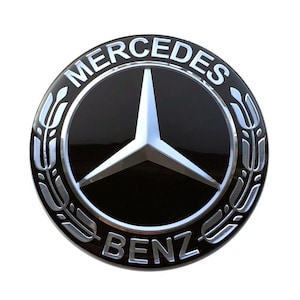 May include: A circular black and silver emblem featuring the Mercedes-Benz three-pointed star logo in the center. The word MERCEDES is arched across the top and BENZ is arched across the bottom, with silver laurel leaf accents on the sides.