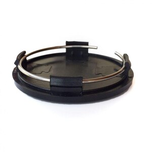 May include: A circular black plastic wheel center cap featuring a smooth, flat top surface and a series of black mounting clips along the underside. A thin, silver-toned metal tension ring is fitted around the inner perimeter of the clips.
