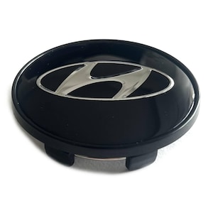 May include: A circular black wheel center cap featuring a silver Hyundai logo in the center. The cap has a glossy finish on the top surface and includes black plastic mounting clips on the underside for secure attachment to a vehicle wheel.