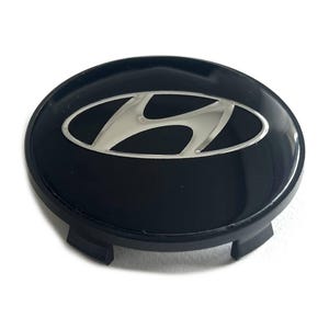 May include: A circular black wheel center cap featuring a silver Hyundai logo emblem in the center. The cap has a glossy finish on the top surface and includes black plastic mounting clips on the underside for secure attachment to a vehicle wheel.