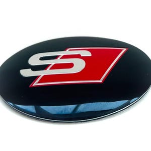 May include: An oval-shaped emblem featuring a glossy black background with a central red parallelogram. A metallic silver letter S is positioned over the red shape, with a thin silver border outlining the red parallelogram and the entire oval emblem.