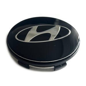 Könnte beinhalten: Eine kreisförmige, schwarze Radnabenabdeckung mit einem silbernen Hyundai-Logo in der Mitte. Die Abdeckung hat eine glänzende Oberfläche und eine schwarze Kunststoffbasis mit sichtbaren Befestigungsclips am unteren Rand für die Radmontage.