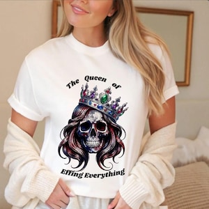 Może przedstawiać: Biały t-shirt z krótkim rękawem z grafiką czaszki z długimi włosami w koronie wysadzanej klejnotami. Tekst nad czaszką brzmi The Queen of, a tekst poniżej brzmi Effing Everything, zapisany pogrubioną czcionką w kolorze czarnym.