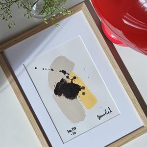 Puede incluir: Una pintura abstracta enmarcada sobre papel blanco con una pincelada gris claro, una pincelada amarilla y una mancha de pintura negra central con salpicaduras de tinta negra. El texto No. 128-26 está impreso en negro en la parte inferior izquierda.