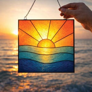Pode incluir: Um apanhador de sol quadrado em vitral apresentando uma cena vibrante de pôr do sol com um sol amarelo brilhante, raios laranja e ondas oceânicas azuis em camadas. A peça é emoldurada em metal preto e inclui uma corrente prateada.