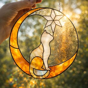 Puede incluir: Un atrapasoles circular de vidrieras que presenta una luna creciente naranja y un gato blanco sentado en la curva. El gato se estira hacia una estrella de vidrio transparente y texturizada en la parte superior. Tiene líneas de plomo negras.
