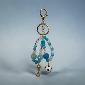 May include: A keychain featuring a gold-toned lobster clasp and ring attached to two loops of blue, white, and translucent beads. One loop includes white letter beads spelling #10, while the other features a gold trophy charm and a soccer ball charm.