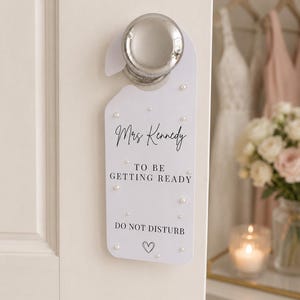 May include: A white rectangular door hanger with rounded corners, featuring the black printed text "Mrs Kennedy TO BE GETTING READY DO NOT DISTURB" and a small heart icon. The surface is decorated with scattered small white pearl accents.