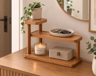 May include: A two-tiered wooden display shelf with rounded corners. The top tier holds a speckled ceramic pot with a trailing green plant. The middle tier holds a ribbed white ceramic dish with keys. The bottom tier holds a candle and a fabric box.