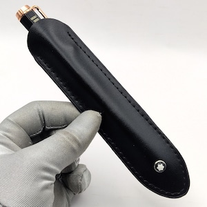 Könnte beinhalten: Eine schwarze Stiftehülle aus Leder mit sichtbaren Nähten an den Kanten und einem weißen Sternemblem am unteren Ende. Ein Stift mit roségoldener Kappe und schwarzem Schaft ist teilweise eingesteckt, wobei der Text 6836/9000 am Clip zu sehen ist.