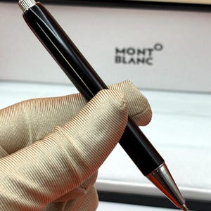 Pode incluir: Uma caneta esferográfica preta com acabamento polido e detalhes em metal prateado na ponta e no topo. A caneta apresenta um corpo cilíndrico elegante e está posicionada na diagonal. O nome da marca Montblanc é visível no fundo branco.