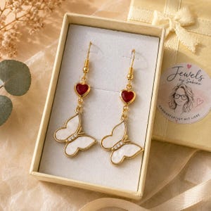 Könnte beinhalten: Ein Paar goldfarbene Ohrhänger mit einem kleinen roten Herzanhänger über einem weißen Schmetterlingsanhänger mit goldenem Rand und kleinen klaren Kristallen am Schmetterlingskörper, präsentiert in einer beigen rechteckigen Geschenkbox mit Aufkleber.