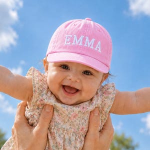 May include: A pink baseball cap featuring the name EMMA embroidered in white capital letters on the front panel. The cap has a curved brim and a structured crown, designed with a classic six-panel construction and a button on the top center.