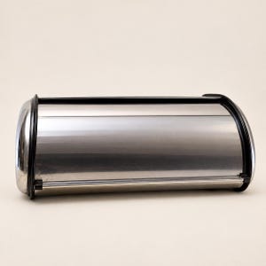 May include: A horizontal, cylindrical bread box featuring a polished, reflective stainless steel body. The container is framed by two black plastic end caps and includes a matching black handle along the front edge for opening the roll-top lid.
