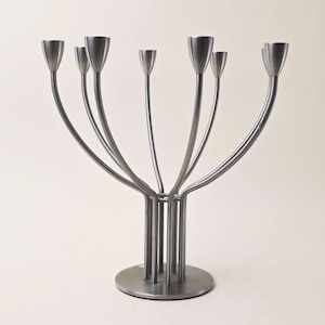 May include: A silver-toned metal candelabra featuring seven individual candle holders supported by slender, curved stems that rise from a central cylindrical base attached to a flat, circular plate. The structure has a matte finish.