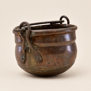 May include: A small, round, antique copper pot with a weathered, dark brown patina. It features a thick, curved metal handle attached to the rim with two riveted, looped brackets on each side. The surface shows signs of oxidation and age.
