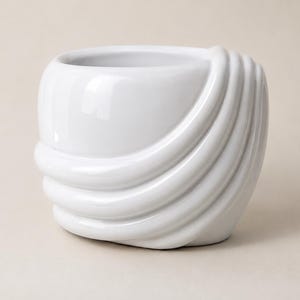 Peut inclure: Un cache-pot en céramique blanc brillant de forme arrondie, semblable à un bol. L'extérieur présente une série de quatre nervures courbes en relief qui s'enroulent sur le côté, créant une texture superposée avec une finition lisse.