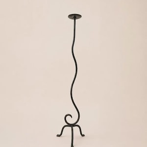 May include: A tall, slender black metal candle holder featuring a single wavy, serpentine stem that rises from a tripod base with curled feet. The top of the holder consists of a flat, circular plate designed to support a candle.