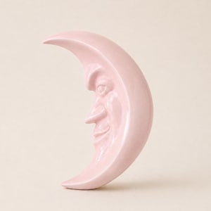 Puede incluir: Una figura de cerámica en forma de media luna de color rosa pálido que presenta un rostro esculpido con una nariz prominente, ojos cerrados y una sonrisa sutil. El objeto tiene un acabado liso y brillante, apoyado verticalmente sobre su punta.