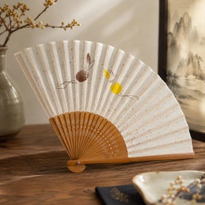 May include: A folding hand fan with a white paper leaf featuring gold flecks and a minimalist illustration of a brown rabbit and a yellow moon. The fan has a light brown bamboo frame with intricate cut-out patterns at the base of the ribs.