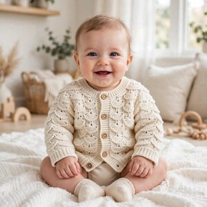 May include: A cream-coloured long-sleeve cardigan featuring a textured fan-shaped knit pattern and five small wooden buttons down the front. The garment has a round neckline and ribbed cuffs, providing a classic look for an infant's wardrobe.