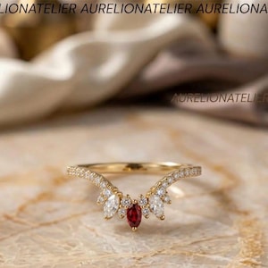 May include: A gold V-shaped ring featuring a central marquise-cut red gemstone flanked by two marquise-cut clear stones and smaller round clear stones along the band. The text AURELIONATELIER is visible in the upper and middle sections of the image.