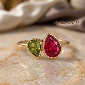 Peut inclure: Une bague en or avec deux pierres précieuses en forme de poire serties clos. Une pierre est un péridot vert vif et la plus grande est un rubis rouge profond. L'anneau est fin et en or poli. Le texte AURELIONATELIER est visible sur l'image.