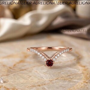 May include: A rose gold V-shaped ring featuring a central round-cut dark red garnet gemstone. The band is accented with a series of small, clear pavé-set diamonds along both sides of the chevron design. The text AURELIONATELIER is visible.
