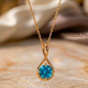 Pode incluir: Um colar de tom dourado com uma corrente delicada e um pingente em forma de gota torcida. O pingente contém uma única pedra preciosa redonda de cor azul brilhante no centro. O texto AURELIONA é visível no fundo.