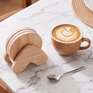 Puede incluir: Un juego de posavasos redondos de madera clara sujetos en un soporte de madera en forma de V, colocados junto a una taza de cerámica color canela con un café latte con arte de espuma en forma de corazón y una pequeña cuchara de metal plateado.