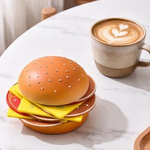 May include: A stackable burger-shaped coaster set featuring a tan bun top with white sesame seeds, yellow cheese slices, red tomato slices, and a brown bottom bun, accompanied by a ceramic mug filled with coffee featuring heart-shaped latte art.