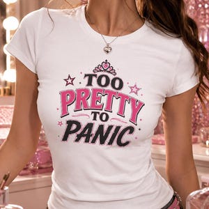 May include: A white short-sleeved t-shirt featuring a graphic print with a pink tiara icon and the text "TOO PRETTY TO PANIC" in bold pink and black lettering, accented with small pink stars on the front chest area of the garment.