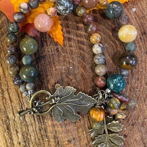 Op de afbeelding: Een meerstrengs kralenarmband met diverse ronde, gepolijste natuurstenen in tinten groen, bruin, beige en roze. De armband is voorzien van een bronskleurige toggle-sluiting in de vorm van een blad en een bungelende, bladvormige bedel.