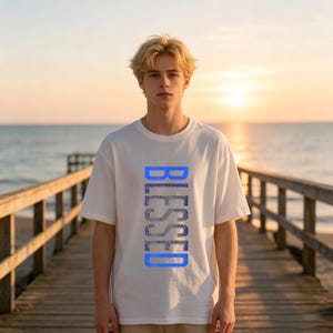 May include: A white short-sleeved t-shirt featuring the word BLESSED printed vertically in a bright blue, textured font across the center of the chest. The garment has a crew neckline and a relaxed fit, constructed from a solid white fabric.
