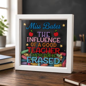 May include: A square white shadow box frame containing a black background with colourful text that reads: Miss Bates THE INFLUENCE OF A GOOD TEACHER CAN NEVER BE ERASED. The frame is filled with various pastel-coloured chalk pieces at the bottom.