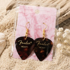 Puede incluir: Un par de pendientes colgantes hechos con púas de guitarra Fender Heavy con patrón de carey marrón. Las púas tienen el texto dorado Fender y HEAVY. Los pendientes están sujetos a una tarjeta con patrón de mármol rosa en una bolsa.