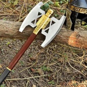 Peut inclure: Une hache de combat à double tranchant avec une tête en métal argenté poli dotée de découpes triangulaires, un connecteur central doré et un long manche en bois avec une base noire. Un casque en métal noir et doré est placé à proximité.