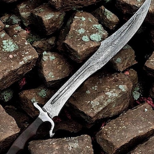 May include: A long, curved sword featuring a distinct Damascus steel blade with a complex, wavy pattern. The hilt is crafted from dark wood and includes a metallic guard with curved quillons, leading to a pommel at the base of the handle.