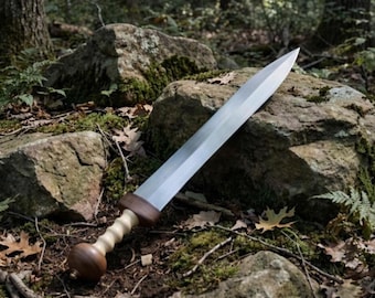 Puede incluir: Una espada de doble filo con una hoja de acero larga y puntiaguda. La empuñadura presenta un pomo y una guarda de madera marrón oscura, con un mango de madera estriada de color claro entre ambos, apoyada sobre una roca cubierta de musgo.