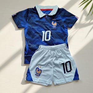 Puede incluir: Una camiseta de f&uacute;tbol azul oscuro con cuello azul claro, rayas negras diagonales y el n&uacute;mero 10 en blanco. Presenta un emblema de gallo dorado con dos estrellas y el texto FFF. Combinada con pantalones cortos azul claro con el n&uacute;mero 10 en negro.