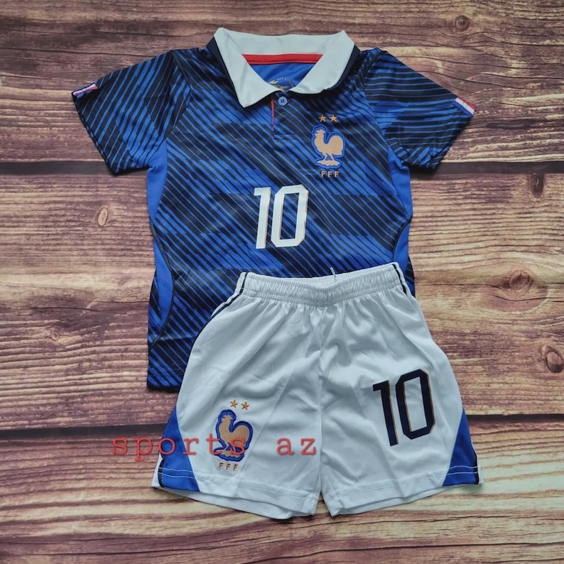 Puede incluir: Una camiseta de f&uacute;tbol a rayas azules y negras con cuello blanco, el n&uacute;mero 10 y el escudo de la selecci&oacute;n francesa, junto con un pantal&oacute;n corto de f&uacute;tbol blanco con el n&uacute;mero 10, ribete azul y el escudo de la selecci&oacute;n francesa en la pierna.