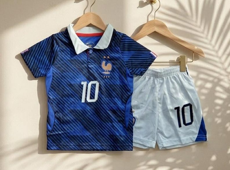 Puede incluir: Una camiseta de f&uacute;tbol azul con cuello blanco, rayas diagonales, el n&uacute;mero 10 y un emblema de gallo con dos estrellas, junto con pantalones cortos de f&uacute;tbol blancos con el n&uacute;mero 10 en azul oscuro y un panel lateral azul en la pierna.