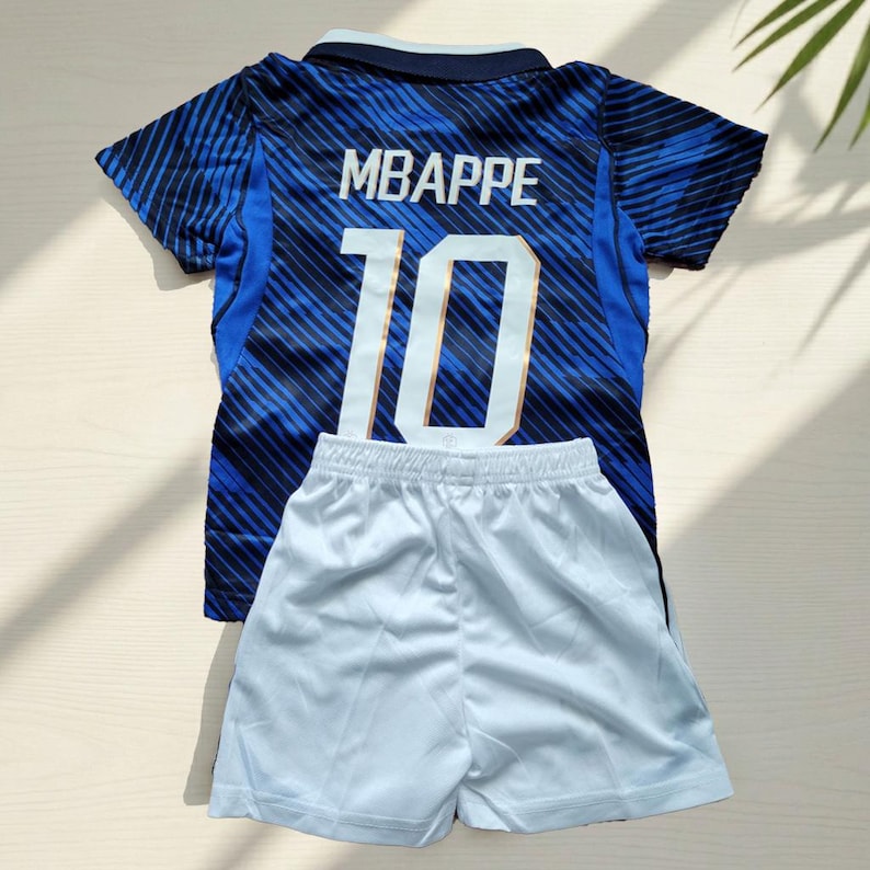Puede incluir: Una camiseta de manga corta azul y azul oscuro con rayas diagonales, que presenta el nombre MBAPPE y el n&uacute;mero 10 en blanco en la parte posterior, combinada con pantalones cortos deportivos blancos lisos. La camiseta tiene un cuello azul oscuro.