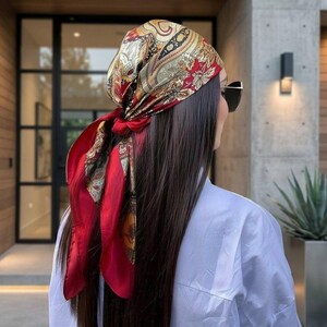Può includere: Un foulard quadrato annodato sulla nuca, caratterizzato da una base rosso vivace con intricati motivi paisley e floreali nei toni dell'oro, del nero e del crema. Il tessuto è ripiegato per coprire la testa, con le estremità annodate saldamente.