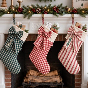May include: Three Christmas stockings hanging from a white fireplace mantel. The left stocking is green and white gingham with the name Oliver, the middle is red and white gingham with the name Evie, and the right is red and white striped with the name Amelia.